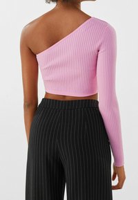 Top corto asimétrico de tejido de canalé en color rosa, con una manga larga y un hombro al descubierto. Combinado con pantalones negros a rayas.