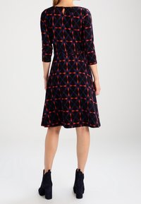 Kvinna i en knälång svart klänning med röda och blå geometriska mönster, trekvartslånga ärmar och svarta klackade ankelskor, sedd bakifrån.