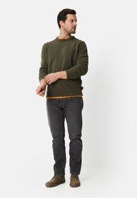 Mørkegrøn strikket sweater med ribbede manchetter og kant, lag på en sennepsgul skjorte. Båret med mørkegrå jeans og brune sko.
