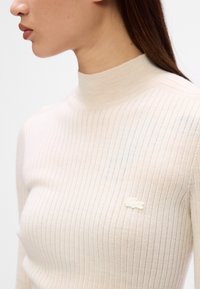 Top de gola alta canelado em bege claro, feito de material suave, apresentando um pequeno logótipo branco da Lacoste no peito. Design liso e ajustado.