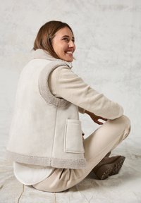 Beige ärmellose Weste mit einem weichen, strukturierten Kragen, Fronttasche und glatter Oberfläche; getragen über einem hellen Pullover und beigen Hosen.
