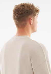 Tricou oversized de culoare bej deschis, cu guler rotund și design cu umeri lăsați, fiind confecționat din material de bumbac moale și având o textură netedă.