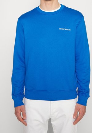 Mężczyzna ubrany w jasnoniebieską bluzę Emporio Armani z białym logo na piersi i białe spodnie, stojący na jednolitym tle.