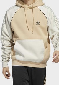 Beige hoodie med vita accenter, ribbade ärmslut och en framficka. Har svart adidas-logga och kontrasterande linjedetaljer på ärmarna.
