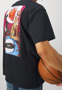 Camiseta de algodón negra con un gráfico colorido en la parte trasera que presenta un leopardo dorado, balones de baloncesto y la marca Nike.