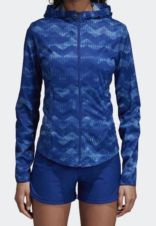 Veste zippée bleue avec capuche, présentant un motif en chevrons et un tissu texturé. Associée à un short bleu assorti.