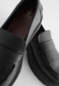 Black leather loafers with a textured finish, featuring a fringed design and a thick black sole. Inside has a branded brown insole.