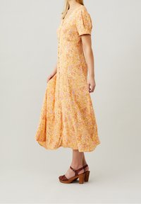 Vestido midi amarillo con flores, mangas abullonadas, detalle de botones y una silueta fluida. Combinado con sandalias de plataforma marrones.