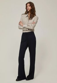 Donna in piedi con le braccia incrociate, vestita con una camicia a righe beige e bottoni, pantaloni neri a gamba larga e scarpe nere, su uno sfondo neutro.