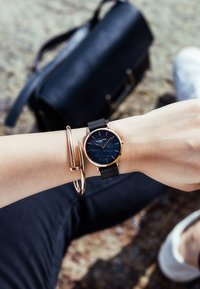 Reloj con correa de malla negra, de caja redonda en tono dorado y esfera azul oscura. Acompañado de una pulsera dorada. Bolsa negra en el fondo.