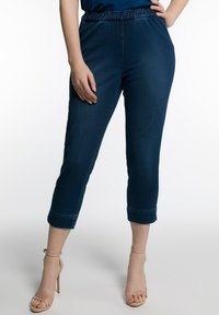 Pantalones ajustados azul oscuro con una cinturilla elástica y dobladillos con puños, fabricados en tela de mezclilla. Llevados con tacones abiertos nude.