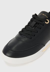 Black leather sneaker featuring contrasting white rubber sole, textured black laces, and gold-tone accents, including a logo tag.
