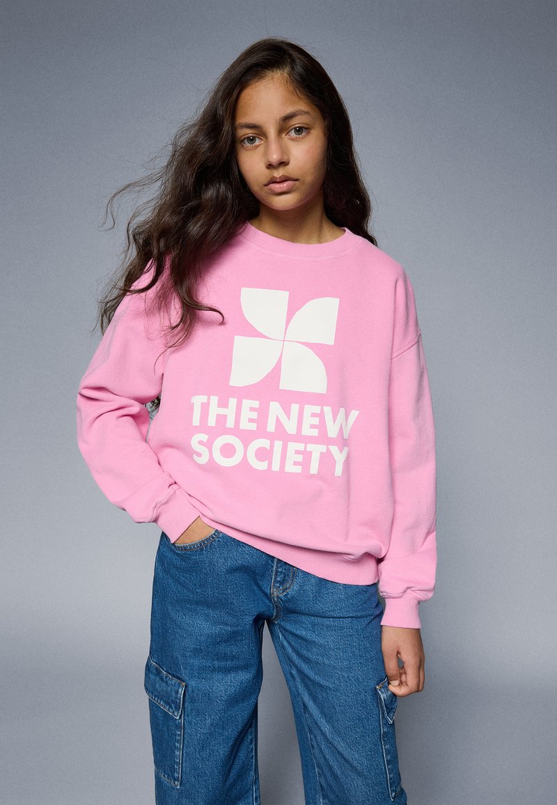 Roze sweatshirt met een wit logo met een geometrisch ontwerp en tekst die leest "DE NIEUWE SAMENLEVING," gecombineerd met blauwe cargo-broek.