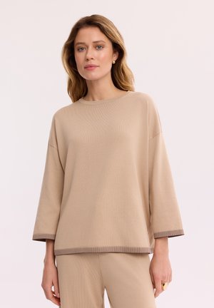 Kobieta o blond włosach do ramion, ubrana w beżową, luźną bluzkę i spodnie, stojąca na tle jednolitego jasnego tła.