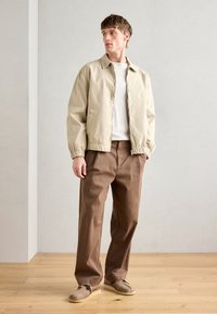 Beige pamut dzseki, rugalmas mandzsettával, barna nadrággal párosítva. A modell egy fa padlón áll, amely bemutatja az öltözetet egy semleges környezetben.