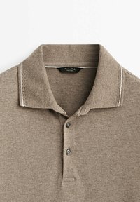 Polo shirt marron avec un tissu texturé. Il présente un col, deux boutons et des coutures blanches contrastantes sur le col.