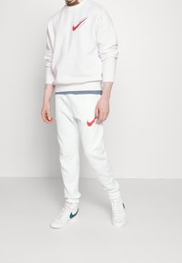 Sudadera blanca de Nike con logo rojo, combinada con pantalones de chándal blancos que presentan un logo en acento rojo. Modelo con zapatillas blancas que tienen el swoosh verde de Nike.