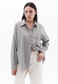 Camicia a righe grigia e bianca, con maniche lunghe, colletto a punta, tasca sul petto e vestibilità morbida. Indossata con pantaloni beige.