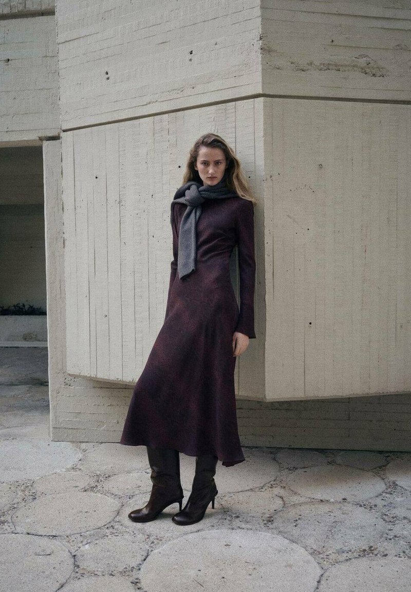 Bordeauxrode jurk met lange mouwen, een aansluitend lijfje en een vloeiende rok, gecombineerd met een grijze sjaal en bruine laarzen met hak. Stoffen afwerking met textuur.