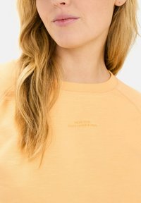 Lys orange sweatshirt med ribbet halsudskæring, med prægede bogstaver "LAVET TIL DAGLIGE EVENTYR" på forsiden, og tekstureret stof.