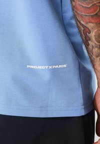 Camiseta de algodón de color azul claro con cuello redondo, que presenta un logo impreso en blanco "PROJECT X PARIS" en la parte inferior izquierda.
