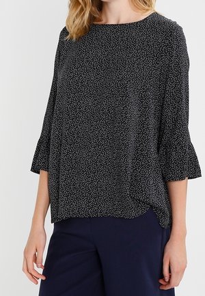 Woman wearing a loose black blouse with small white dots and three-quarter sleeves paired with dark blue pants.