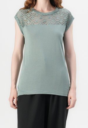 Top verde claro con mangas cortas, que presenta un panel de encaje transparente con un patrón de ondas en los hombros y una tela suave y ligera.