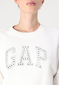 Felpa bianca con la scritta "GAP" adornata con borchie argentate. Presenta un collo rotondo e una texture morbida. Design semplice e contemporaneo.