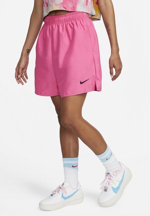 Personne portant un short Nike rose vif, des baskets blanches Nike avec des swooshes bleus, des chaussettes blanches à rayures bleues et rouges, debout sur un fond blanc.