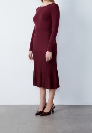 Bordeauxrote, gerippte Strickkleid mit tailliertem Oberteil und ausgestelltem Saum, kombiniert mit glänzenden bordeauxroten spitzen Pumps.