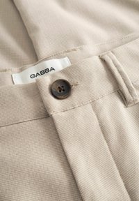 Heleda beežikat tekstuuriga kangast püksid tumeda nelja augu nupuga ja nähtava "GABBA" brändisildiga vöökohal.