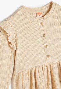 Beżowa, prążkowana sukienka dla niemowląt z okrągłym dekoltem, falbaną na ramionach oraz pięcioma brązowymi guzikami z przodu. Miękka, teksturowana tkanina.