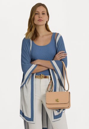 Femme portant un haut bleu sans manches et un châle rayé bleu et blanc avec un pantalon blanc, tenant un sac bandoulière beige avec un logo doré.