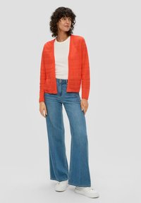 Gilet texturé orange avec une coupe décontractée, porté sur une chemise blanche, associé à un jean en denim à jambes larges et des baskets blanches.