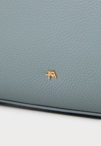 Pilkos akmeninės odos su lygiu paviršiumi, apatinėje kampo dalyje yra auksinė metalinė logotipo detalė.