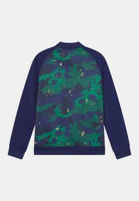 Marinblå idrottsjacka med ett grönt och mörkblått kamouflage-mönster. Har raglanärmar och en ribbad krage.