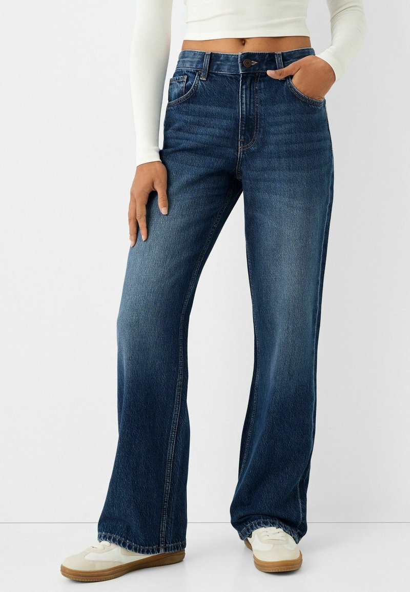Femme portant un jean taille haute bleu foncé à jambes larges, des baskets blanches et un top blanc à manches longues court, main dans la poche.