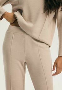 Beżowy zestaw dzianinowy do noszenia w domu, składający się z luźnej bluzki z długim rękawem oraz dopasowanych spodni zwężanych ku dołowi z detalami szwów i miękką fakturą.