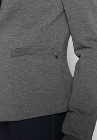 Grå jacka i en texturerad stickning, har en liten sidoficka och en dekorativ detalj i svart metall vid fållen.