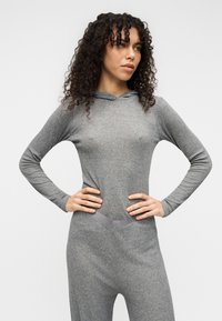 Γκρι φορμάκι με κουκούλα, μακριά μανίκια, απαλή υφή, εφαρμοστή σχεδίαση, διακριτική λεπτομέρεια στη μέση και ελαφρώς στενότερα παντελόνια.