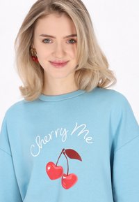 Ljusblå sweatshirt i bomull, med vit "Cherry Me"-tryck och röda körsbär. Avslappnad passform och rund halsringning.