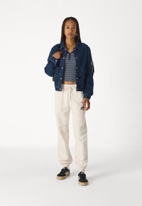 Giacca di jeans, top corto a righe, pantaloni sportivi beige con cordino, sneakers nere con strisce bianche, in piedi su uno sfondo neutro.