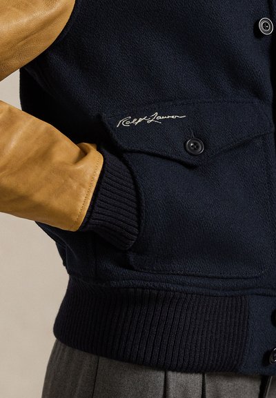 Ναυτικό μπουφάν φτιαγμένο από μαλλί με καφέ δερμάτινα μανίκια, ribbed cuffs και κουμπωτό μπροστινό μέρος, διαθέτοντας υπογραφή κεντήματος στην τσέπη του στήθους.