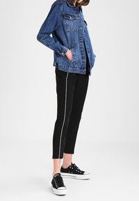 Persona que lleva puesta una chaqueta de mezclilla azul, pantalones negros con franjas laterales blancas y zapatillas negras de caña baja con suelas blancas.