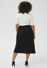 Jupe midi noire avec taille ajustée et fente sur le côté, associée à un haut blanc ajusté. Le tissu semble lisse avec un léger éclat. Talons hauts noirs.
