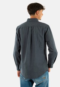 Camisa de manga larga de color gris oscuro con cuello abotonado, pliegue en la parte posterior y dobladillo curvado. Lleva puesta con jeans azules, mostrando un ajuste casual.