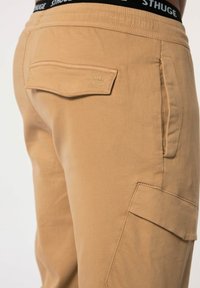 Beige Cargo-Hosen mit einer Gesäßtasche, die ein gesticktes Logo trägt, einem flachen Bund mit kontrastierender Markenkennzeichnung und einer weichen, glatten Stoffstruktur.