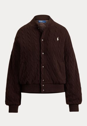 Donna con cardigan marrone scuro a trecce con bottoni, dolcevita grigia, pantaloni larghi grigi, scarpe nere e occhiali da sole scuri, in piedi con le mani in tasca.