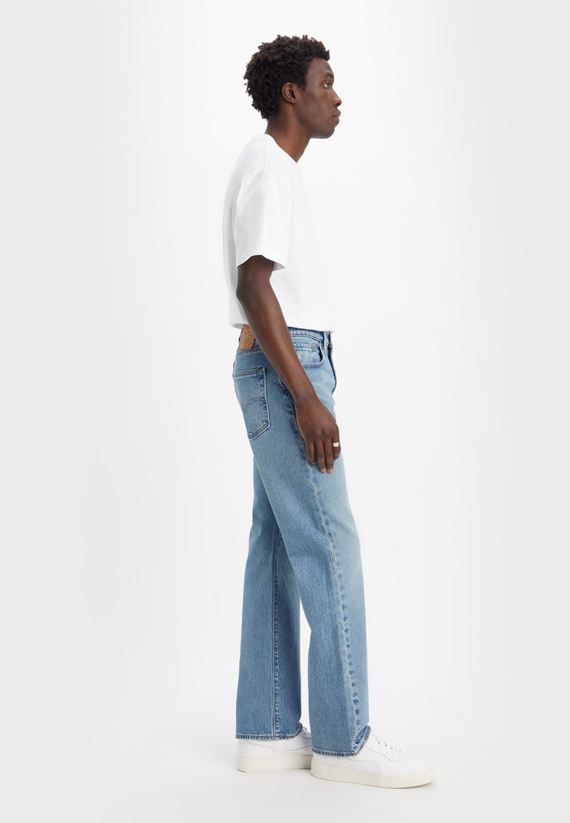 Helle blaue Jeans mit lockerer Passform, mit einem Lederpatch auf der Rückseite, kombiniert mit einem weißen, kurzärmeligen T-Shirt und weißen Sneakers.