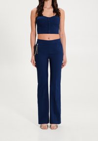 Žena stojící v námořnickém modrém topu bez ramínek a odpovídajících kalhotách s vysokým pasem, s zlatým řetízkovým doplňkem a otevřenou obuví na podpatku.